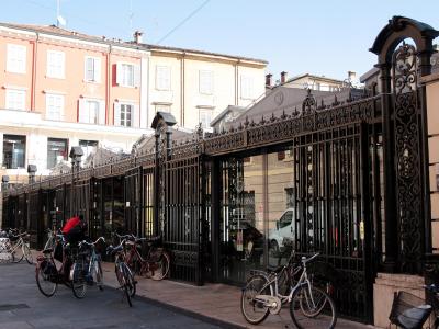 A street-level view of a long, ornate black iron gate and fence enclosing a building with a glass facade, with several bicycles parked on the sidewalk in front and multi-story residential buildings in the background.