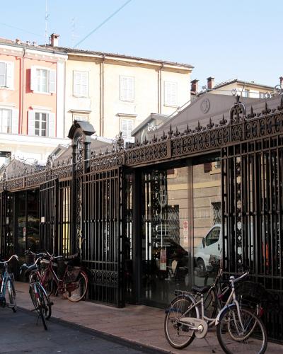 A street-level view of a long, ornate black iron gate and fence enclosing a building with a glass facade, with several bicycles parked on the sidewalk in front and multi-story residential buildings in the background.