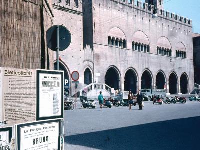A large, historic brick building with a crenellated roofline and a series of pointed arches on the ground floor, situated in a paved public square with parked scooters and posters on a wall in the foreground.