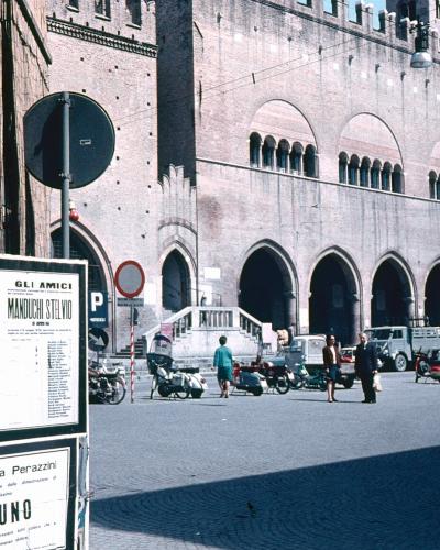 A large, historic brick building with a crenellated roofline and a series of pointed arches on the ground floor, situated in a paved public square with parked scooters and posters on a wall in the foreground.