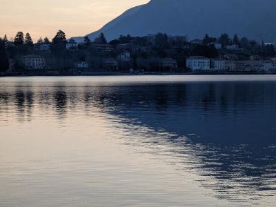 A twilight view across calm water toward a lakeshore town with buildings and trees at the base of a large mountain, with reflections on the water surface.
