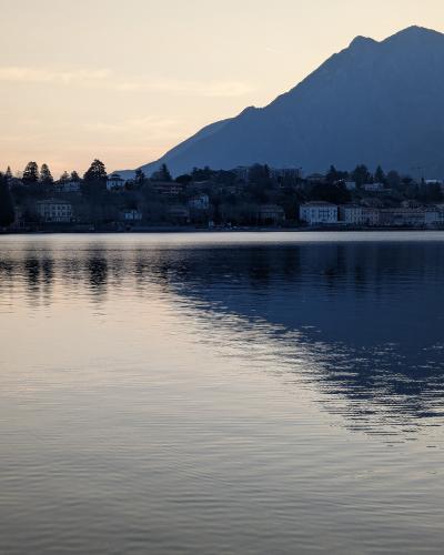 A twilight view across calm water toward a lakeshore town with buildings and trees at the base of a large mountain, with reflections on the water surface.