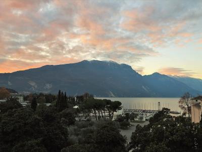 A scenic view of a large lake with a marina and boats in the foreground, surrounded by trees and buildings, with a massive, steep mountain range rising directly behind it under a colorful sunset sky.