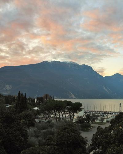 A scenic view of a large lake with a marina and boats in the foreground, surrounded by trees and buildings, with a massive, steep mountain range rising directly behind it under a colorful sunset sky.