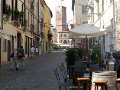 A cobblestone street in an Italian town with outdoor cafe seating in the foreground and a tall medieval brick tower visible at the end of the street, flying a flag; signage for "HOTEL AL CASTELLO" is visible on a building near the tower.