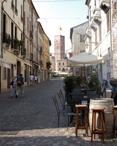 A cobblestone street in an Italian town with outdoor cafe seating in the foreground and a tall medieval brick tower visible at the end of the street, flying a flag; signage for "HOTEL AL CASTELLO" is visible on a building near the tower.
