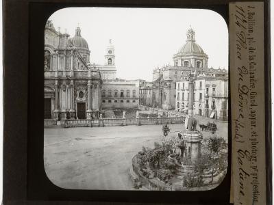 A vintage black-and-white photograph of a large European square featuring a prominent cathedral with a dome and bell tower on the left, a central fountain with an elephant statue in the foreground, and handwritten text on the right border reading