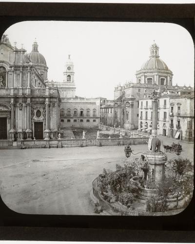 A vintage black-and-white photograph of a large European square featuring a prominent cathedral with a dome and bell tower on the left, a central fountain with an elephant statue in the foreground, and handwritten text on the right border reading