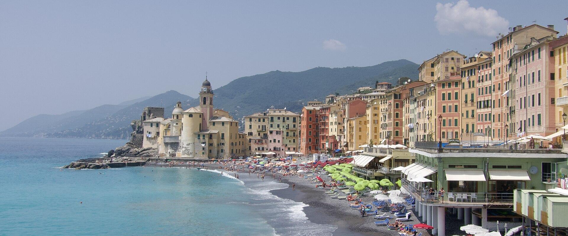 A sunlit coastal scene showing a curved pebble beach lined with colorful umbrellas and beachgoers, backed by a row of pastel-colored buildings and a prominent church with a bell tower on a rocky promontory, with hills rising behind.