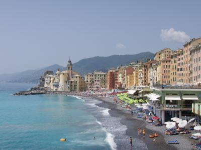 A sunlit coastal scene showing a curved pebble beach lined with colorful umbrellas and beachgoers, backed by a row of pastel-colored buildings and a prominent church with a bell tower on a rocky promontory, with hills rising behind.