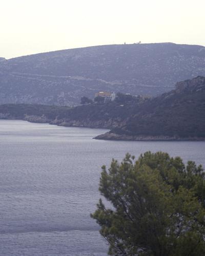 A coastal landscape showing a large, flat-topped limestone headland rising from the sea, with visible structures including a lighthouse on the left and a tower on a lower promontory, framed by green trees in the foreground.