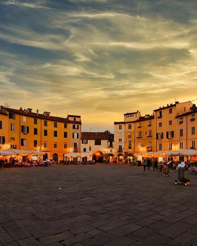 A wide, elliptical cobblestone piazza at dusk, surrounded by multi-story yellow and ochre buildings with shuttered windows and ground-floor cafes with white umbrellas; a person stands near a chalkboard menu in the foreground.