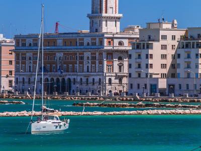 The image shows the Palazzo della Provincia di Bari (Provincial Palace) with its distinctive clock tower, situated directly on the waterfront of Bari, Italy, with a sailboat and a kayaker in the foreground on the Adriatic Sea.