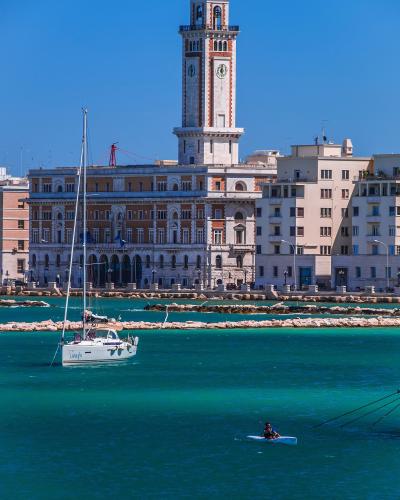 The image shows the Palazzo della Provincia di Bari (Provincial Palace) with its distinctive clock tower, situated directly on the waterfront of Bari, Italy, with a sailboat and a kayaker in the foreground on the Adriatic Sea.