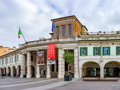 A wide-angle photograph of a grand neoclassical building with a colonnaded portico, featuring an Italian flag and an EU flag, banners advertising "Think big" and a red poster, situated in a paved public square with adjacent buildings including one