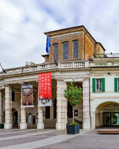 A wide-angle photograph of a grand neoclassical building with a colonnaded portico, featuring an Italian flag and an EU flag, banners advertising "Think big" and a red poster, situated in a paved public square with adjacent buildings including one