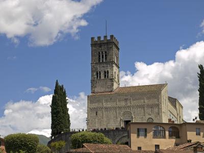 The image shows a large medieval stone church with a prominent square bell tower featuring crenellations and arched openings, set against a blue sky with clouds, with people visible on a terrace in front and snow-capped mountains in the background