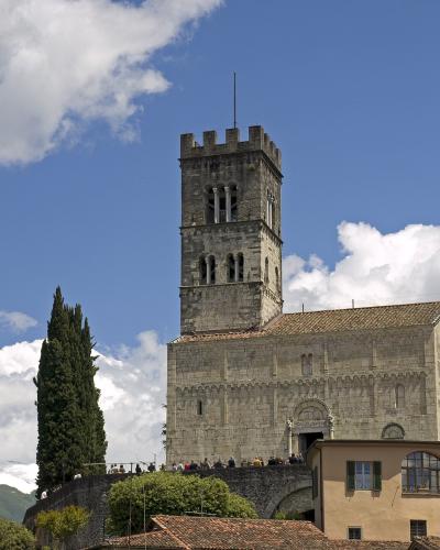 The image shows a large medieval stone church with a prominent square bell tower featuring crenellations and arched openings, set against a blue sky with clouds, with people visible on a terrace in front and snow-capped mountains in the background