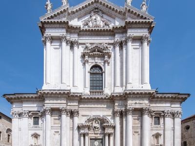 The image shows the white marble Baroque facade of a large cathedral with prominent columns, statues on the roofline, and a central pediment, set against a clear blue sky.