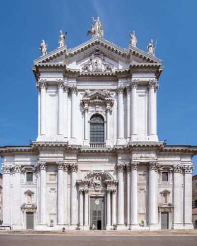 The image shows the white marble Baroque facade of a large cathedral with prominent columns, statues on the roofline, and a central pediment, set against a clear blue sky.
