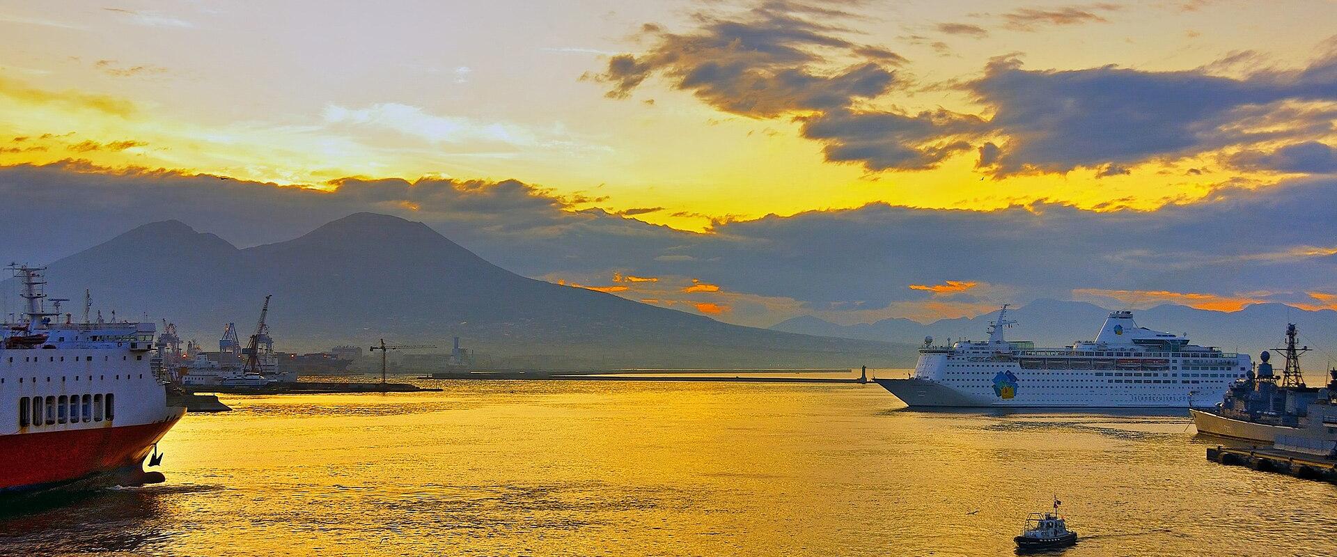 A harbor scene at sunrise or sunset with large ships, including a cruise liner and a red-hulled vessel, on golden water, with a prominent conical mountain (Mount Vesuvius) in the background under a cloudy sky.