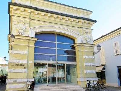 The image shows the exterior of a yellow neoclassical building with large glass windows and doors, labeled "LA PSCARIA 1911" on its facade, with bicycles parked in front and people visible to the left.