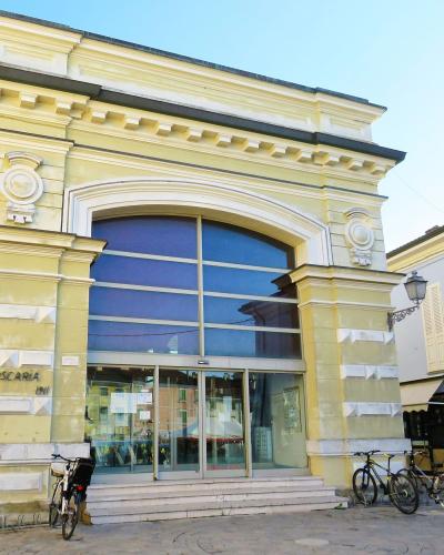 The image shows the exterior of a yellow neoclassical building with large glass windows and doors, labeled "LA PSCARIA 1911" on its facade, with bicycles parked in front and people visible to the left.