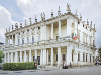 A large, white Renaissance palace with a two-story loggia featuring Doric columns on the ground floor and Ionic columns on the upper floor, topped by statues along the roofline, with Italian and EU flags flying from a balcony.