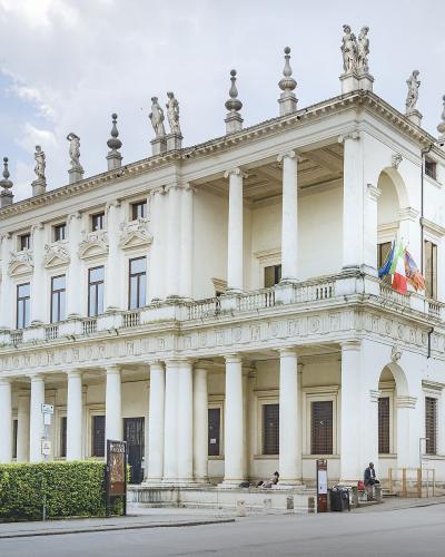A large, white Renaissance palace with a two-story loggia featuring Doric columns on the ground floor and Ionic columns on the upper floor, topped by statues along the roofline, with Italian and EU flags flying from a balcony.