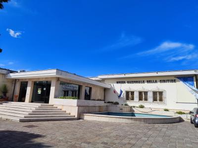 The image shows the exterior of a modern, low-rise white building with a paved courtyard, a small reflecting pool, and prominent signage reading "MUSEO NAZIONALE DELLA SIRITIDE" on the facade, along with Italian and EU flags.