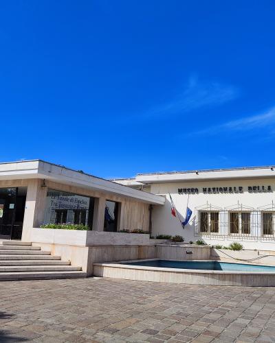 The image shows the exterior of a modern, low-rise white building with a paved courtyard, a small reflecting pool, and prominent signage reading "MUSEO NAZIONALE DELLA SIRITIDE" on the facade, along with Italian and EU flags.