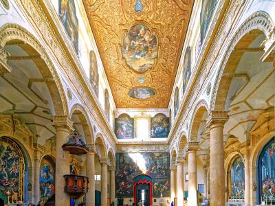 The interior of a grand Baroque cathedral featuring a highly ornate golden ceiling with frescoes, rows of wooden pews, a checkered floor, and large paintings on the walls.