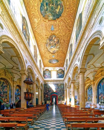 The interior of a grand Baroque cathedral featuring a highly ornate golden ceiling with frescoes, rows of wooden pews, a checkered floor, and large paintings on the walls.