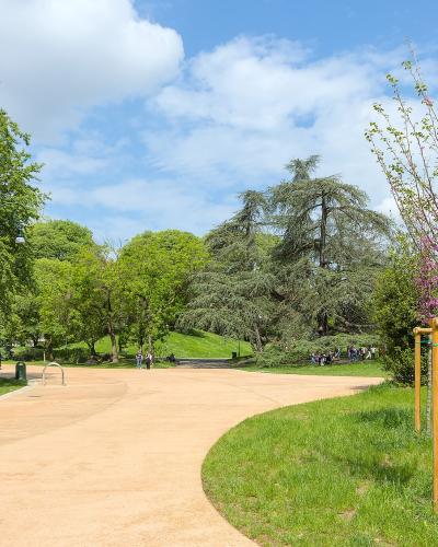 A wide, curving gravel path winds through a lush green park with mature trees, grassy lawns, and a young tree supported by wooden stakes on the right; people are visible in the distance enjoying the space under a partly cloudy sky.