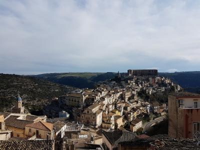 A panoramic view of a historic hillside town in Sicily, featuring dense clusters of ochre and cream-colored buildings with terracotta roofs, prominent Baroque churches with domes and bell towers, and a distinct upper plateau settlement overlooking