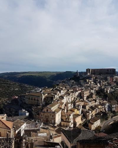 A panoramic view of a historic hillside town in Sicily, featuring dense clusters of ochre and cream-colored buildings with terracotta roofs, prominent Baroque churches with domes and bell towers, and a distinct upper plateau settlement overlooking