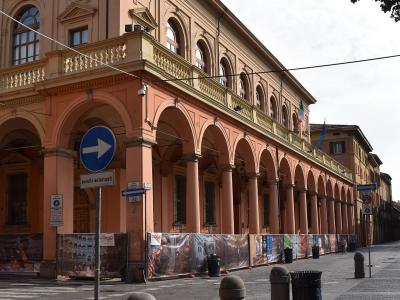 A large, peach-colored historic building with a long ground-floor arcade of arches, featuring Italian street signs (including "eccetto autorizzati"), bollards, and construction hoarding with opera-related imagery along its base.