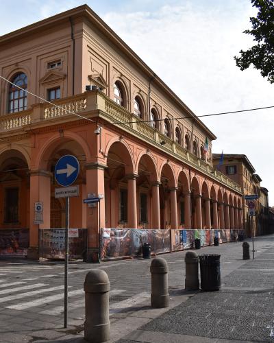 A large, peach-colored historic building with a long ground-floor arcade of arches, featuring Italian street signs (including "eccetto autorizzati"), bollards, and construction hoarding with opera-related imagery along its base.