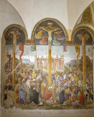 A large, damaged fresco depicting the Crucifixion of Christ flanked by two other crucified figures, with crowds and architectural elements in the background, painted on an interior wall within an arched alcove.