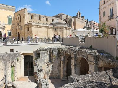 The image shows a sunlit urban scene in Matera, Italy, featuring the ancient rock-cut ruins of the "Palombaro Lungo" cistern in the foreground, with a modern pedestrian walkway and railing above it, and historic buildings including the Church of S