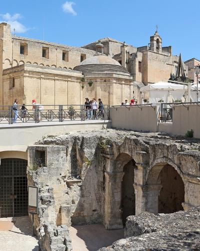 The image shows a sunlit urban scene in Matera, Italy, featuring the ancient rock-cut ruins of the "Palombaro Lungo" cistern in the foreground, with a modern pedestrian walkway and railing above it, and historic buildings including the Church of S