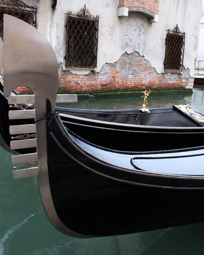 A close-up view of the bow of a traditional black Venetian gondola with its distinctive metal prow ornament (ferro di prua) and golden fittings, floating on a canal next to a weathered building with brickwork and barred windows.