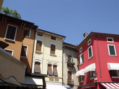 The image shows a street-level view of colorful, multi-story buildings with shuttered windows and awnings over ground-floor shops, under a clear blue sky; visible signage includes “BIANCHI ABBIGLIAMENTO,” “BAR PIZZERIA,” and a parking sign for “CI