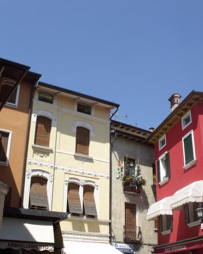 The image shows a street-level view of colorful, multi-story buildings with shuttered windows and awnings over ground-floor shops, under a clear blue sky; visible signage includes “BIANCHI ABBIGLIAMENTO,” “BAR PIZZERIA,” and a parking sign for “CI