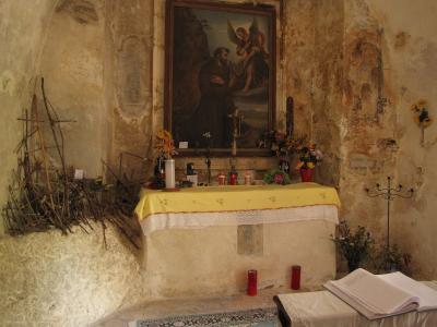 The image shows the interior of a rustic, cave-like chapel or grotto featuring a stone altar with a yellow cloth, a painting of St. Francis of Assisi receiving the stigmata from an angel, and rough-hewn walls with faded frescoes.