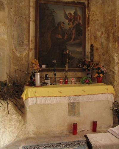 The image shows the interior of a rustic, cave-like chapel or grotto featuring a stone altar with a yellow cloth, a painting of St. Francis of Assisi receiving the stigmata from an angel, and rough-hewn walls with faded frescoes.