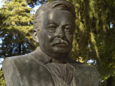 A bronze bust of a man with a mustache, set outdoors against a backdrop of green trees, with the filename indicating it is the "Urbino Busto Pascoli".