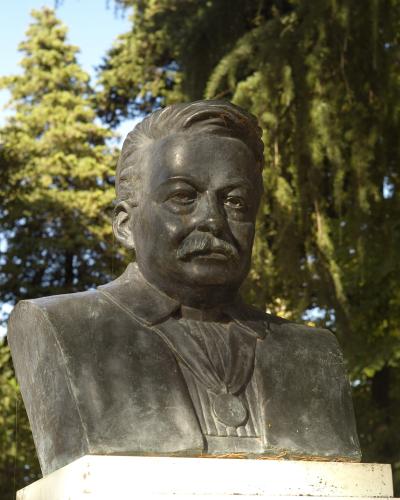 A bronze bust of a man with a mustache, set outdoors against a backdrop of green trees, with the filename indicating it is the "Urbino Busto Pascoli".