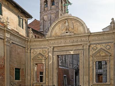 The image shows the ornate marble facade and entrance gate of a Venetian building, featuring a prominent relief of a winged lion (the symbol of St. Mark) and a Latin inscription referencing St. John the Evangelist, with a weathered bell tower risi