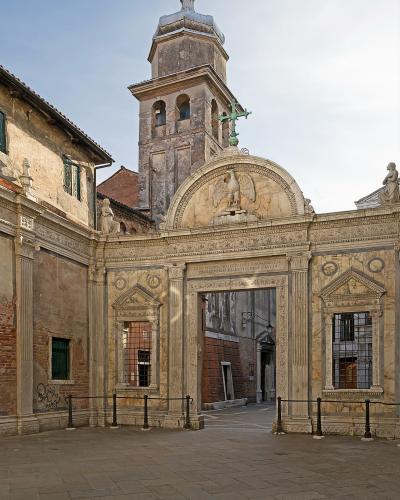 The image shows the ornate marble facade and entrance gate of a Venetian building, featuring a prominent relief of a winged lion (the symbol of St. Mark) and a Latin inscription referencing St. John the Evangelist, with a weathered bell tower risi